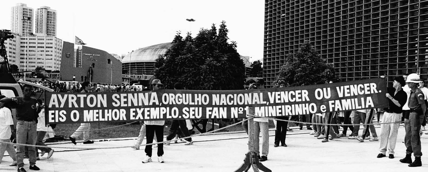 Fans hold up a long hand-painted banner behind a rope barrier on a paved forecourt, a military policeman in a white helmet at one end, tower blocks and a domed arena behind.