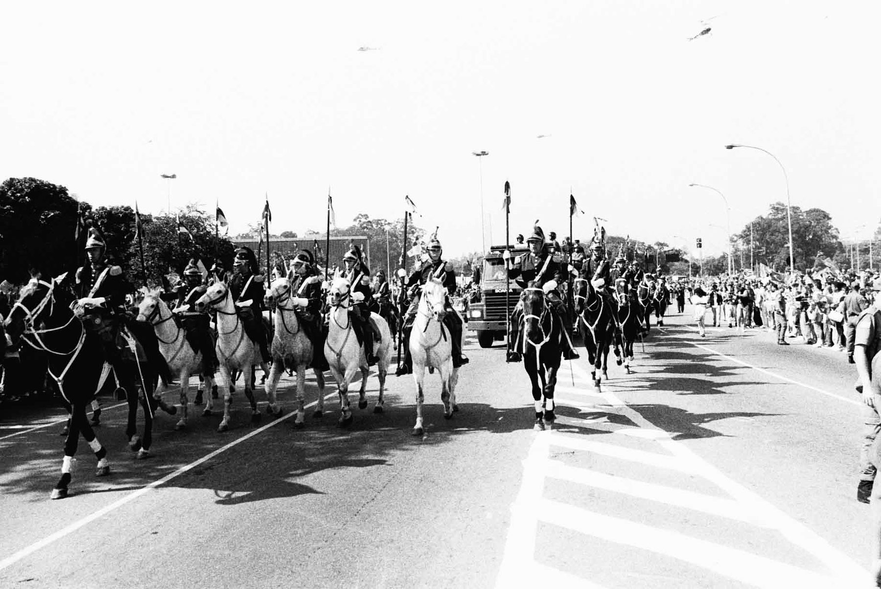A column of mounted lancers on pale and dark horses rides down a broad avenue ahead of a fire engine, spectators lining the road and helicopters in the sky.