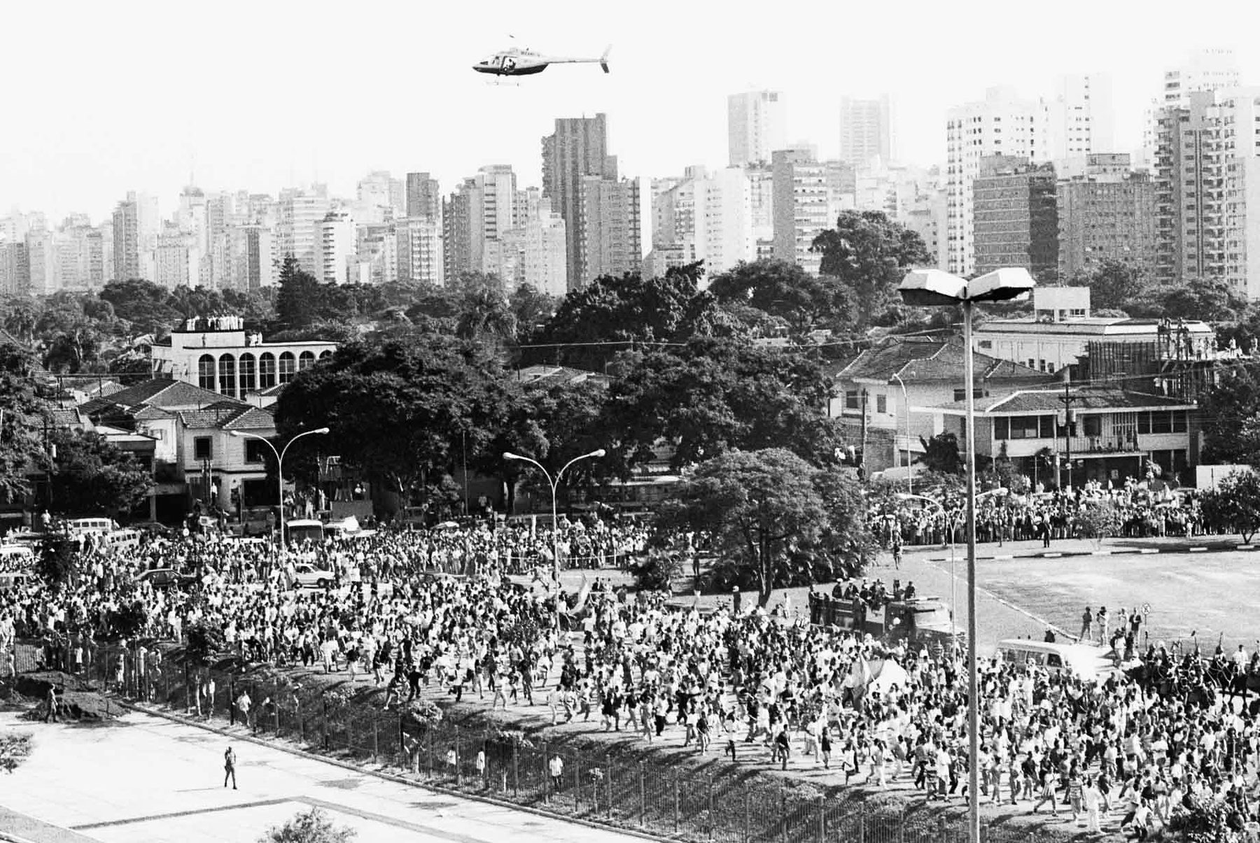 A helicopter hangs in the sky above the tower blocks of São Paulo while a vast crowd fills the road and park below.