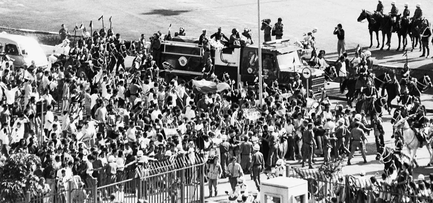 A dense crowd surges around a fire engine with uniformed men standing on top, mounted police beside it and a flag held up in the crowd.