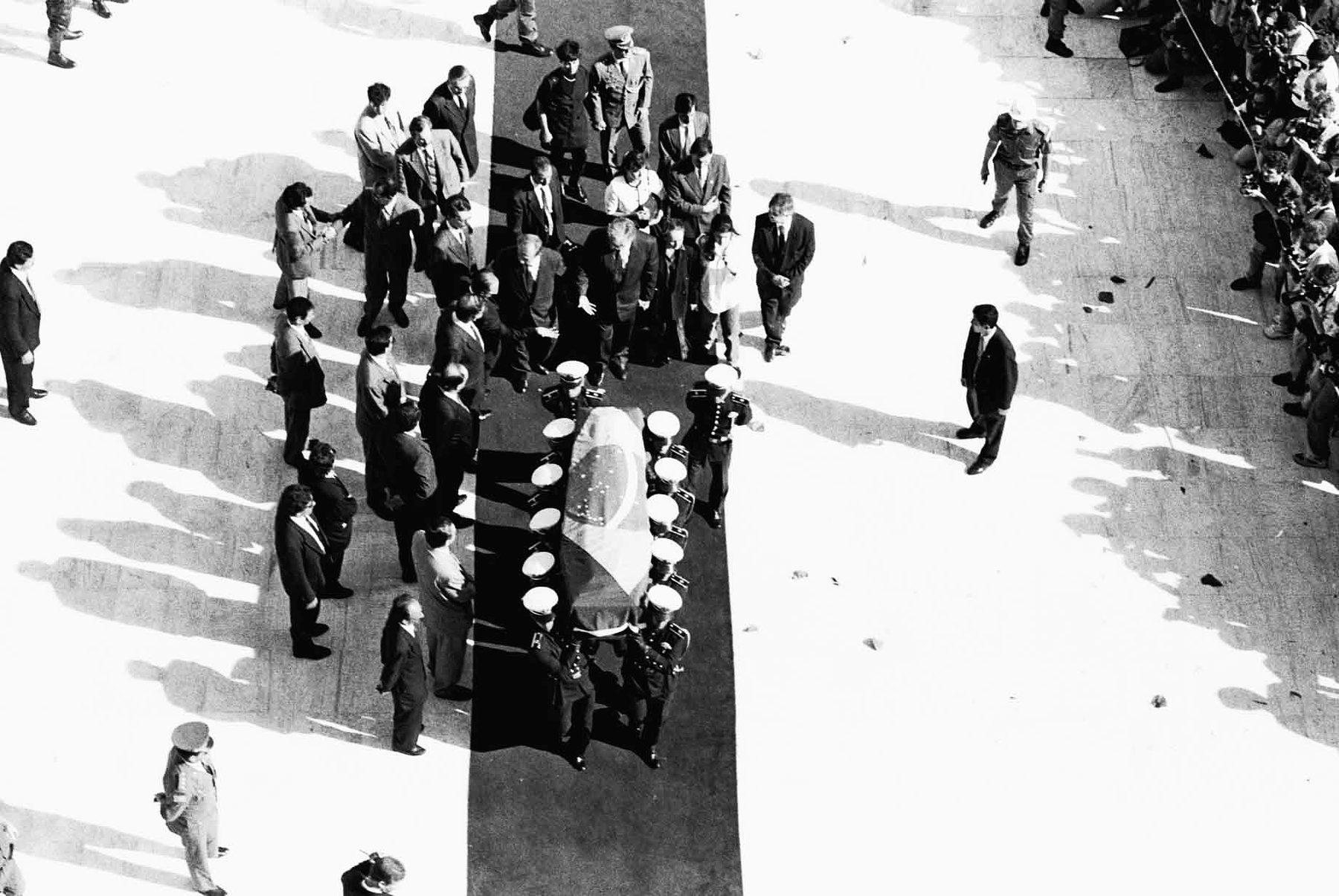 Desde muy arriba, una docena de porteadores de uniforme llevan un ataúd cubierto con una bandera por una alfombra oscura que cruza una explanada soleada; detrás caminan hombres trajeados y los fotógrafos se agolpan en el borde derecho, con largas sombras sobre el pavimento.