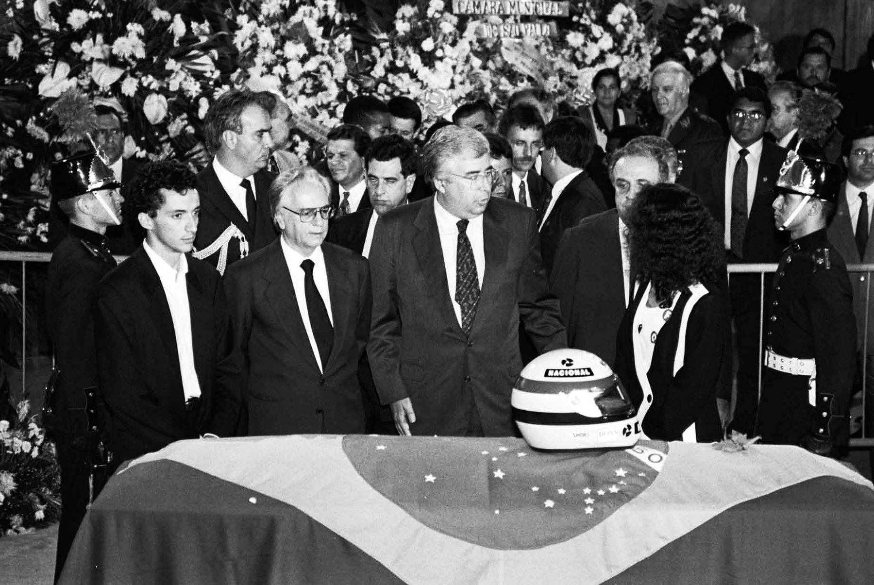 Grey-haired men in dark suits stand with bowed heads at a coffin draped in a flag, a racing helmet resting on it, ceremonial guards in plumed helmets at either side and flowers banked behind.