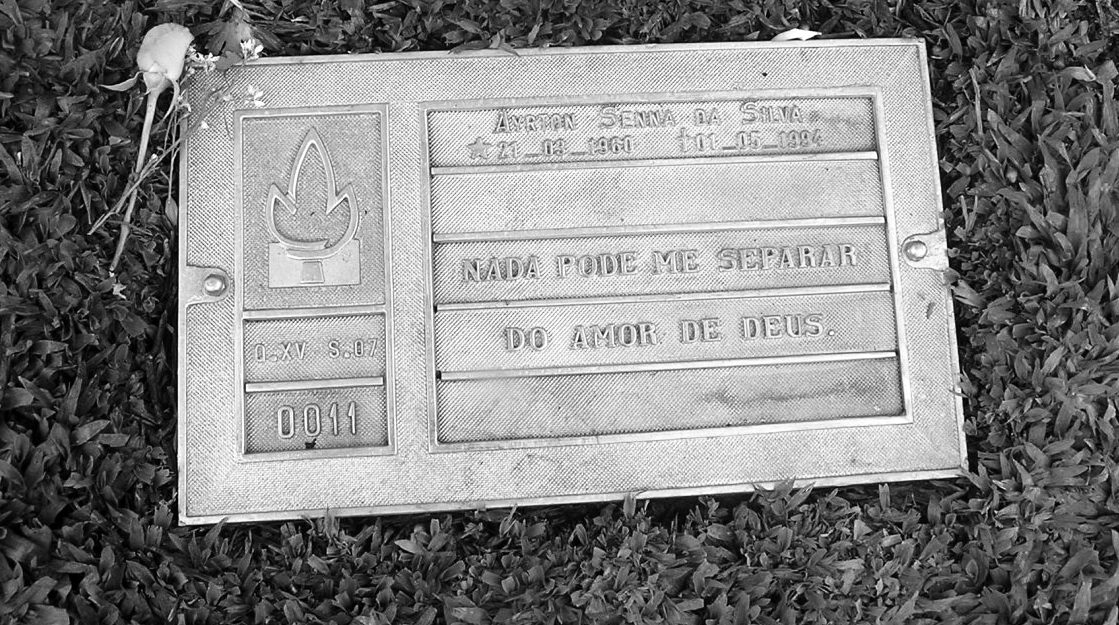 A bronze plaque set in the grass: a flame emblem at the left, his name and dates across the top, two lines of inscription beneath, a rose laid across its top corner.
