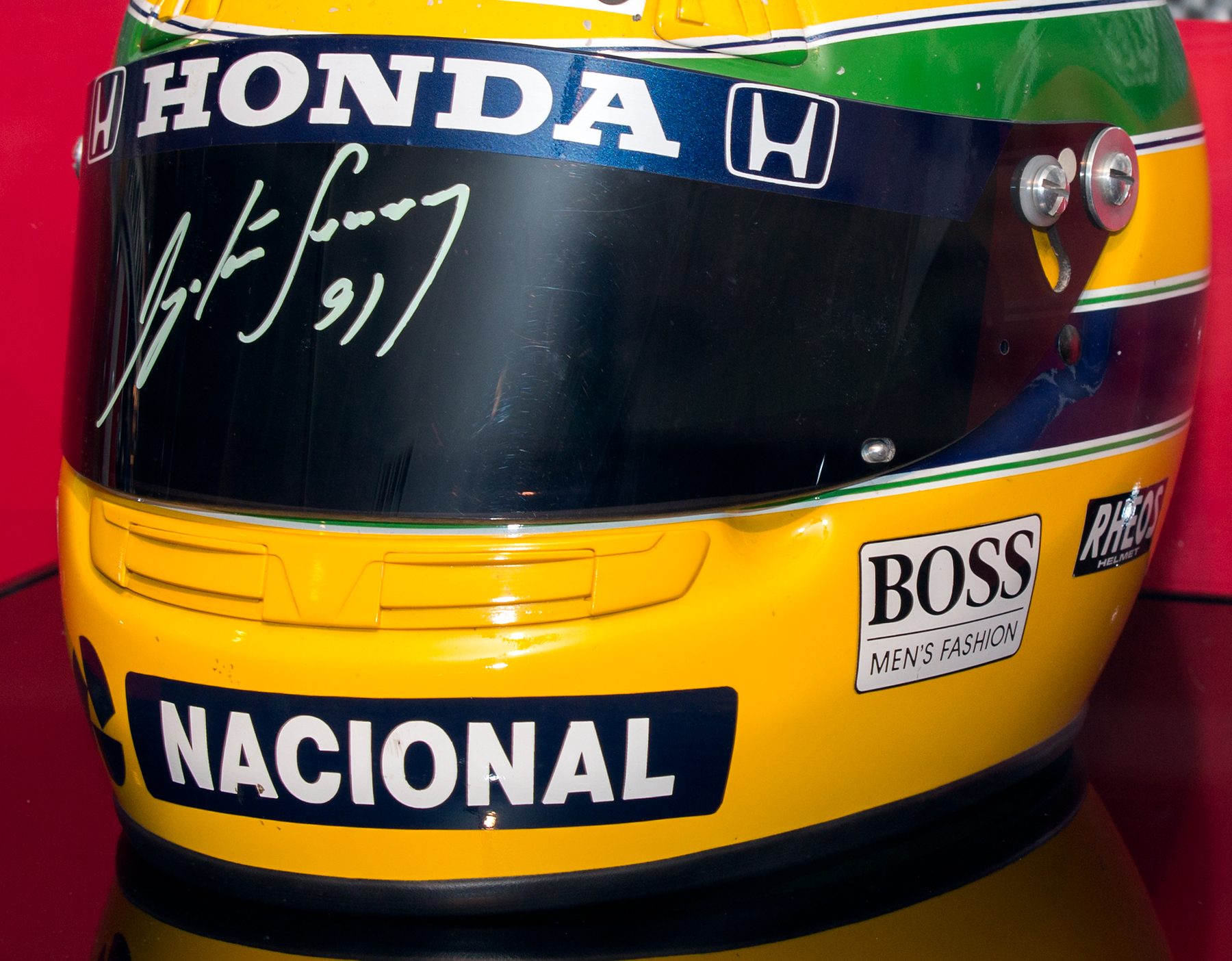 Close-up of a yellow racing helmet with a green band, its dark visor signed in white ink ‘Ayrton Senna 91’ beneath a blue band lettered ‘Honda’, with ‘Nacional’ across the chin.