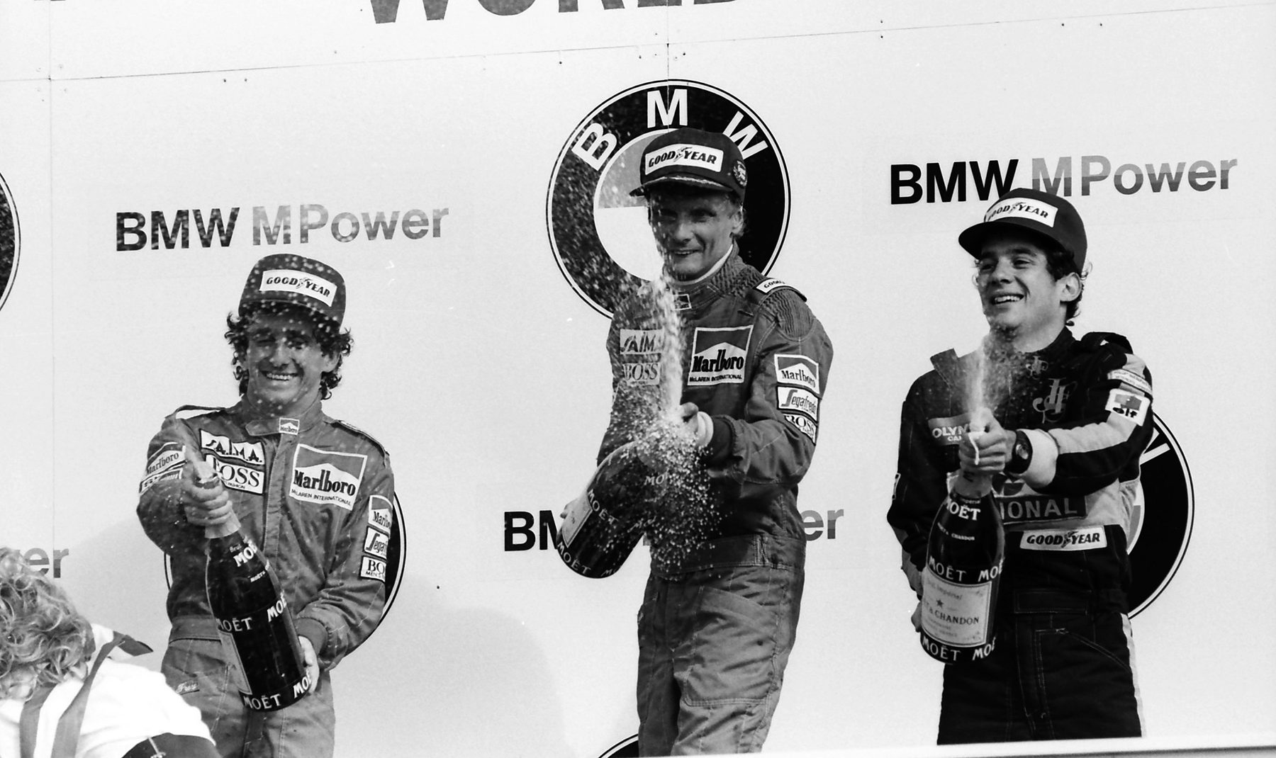 Three drivers in team overalls and caps spray champagne on a podium in front of sponsors’ boards; the youngest, at right, grins broadly with his bottle raised.