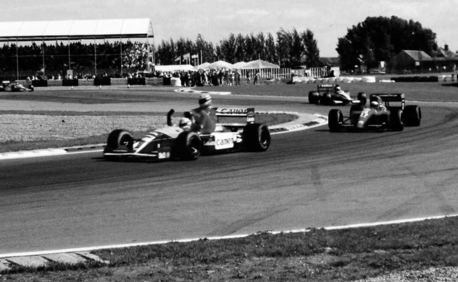 A Williams rounds a curve with a second driver, in dark overalls and a pale helmet, sitting on its sidepod, a darker car following, a grandstand and trees beyond.