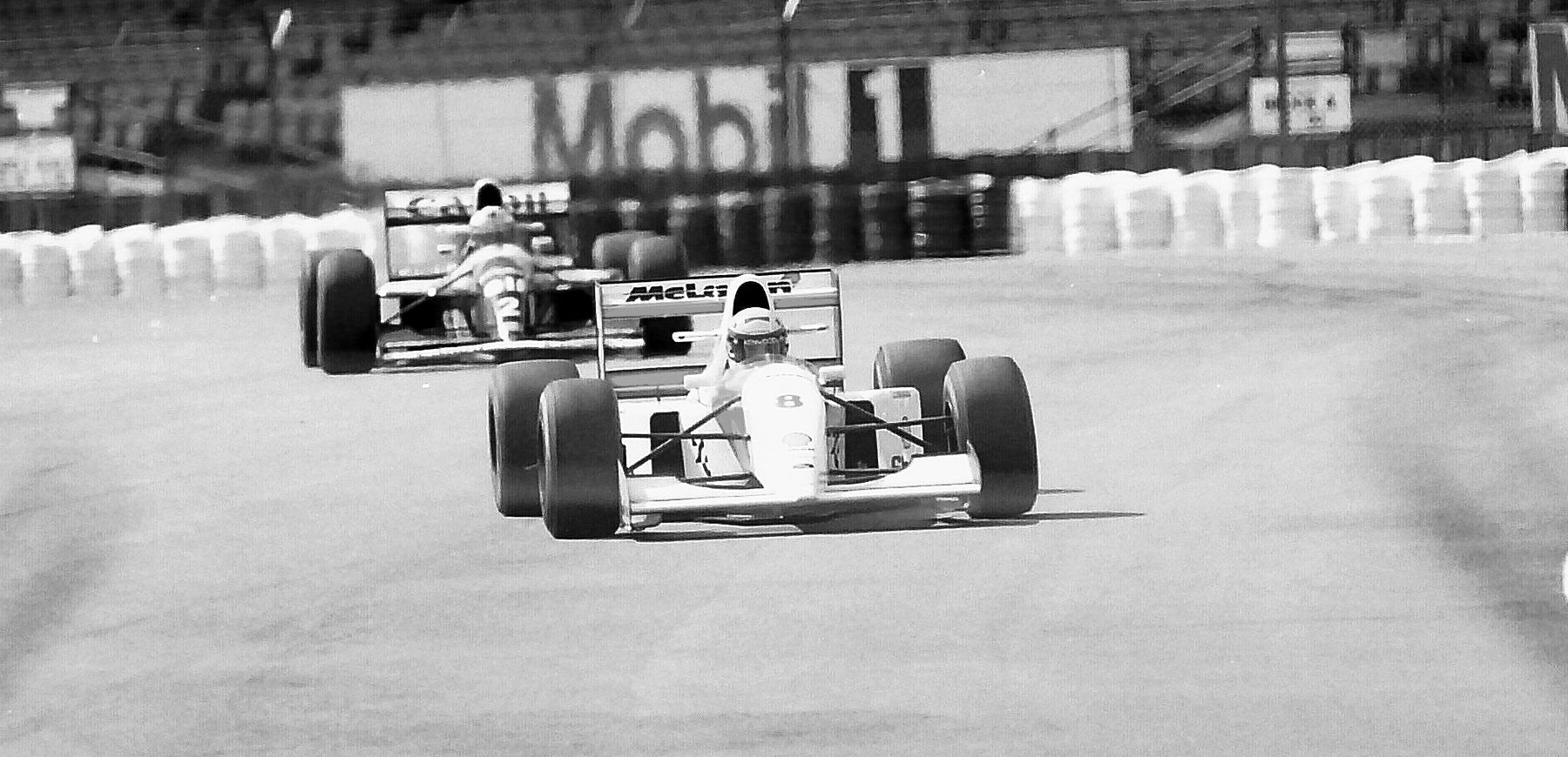 A white McLaren leads a Williams out of a bend on a wide, empty track lined with tyre walls.