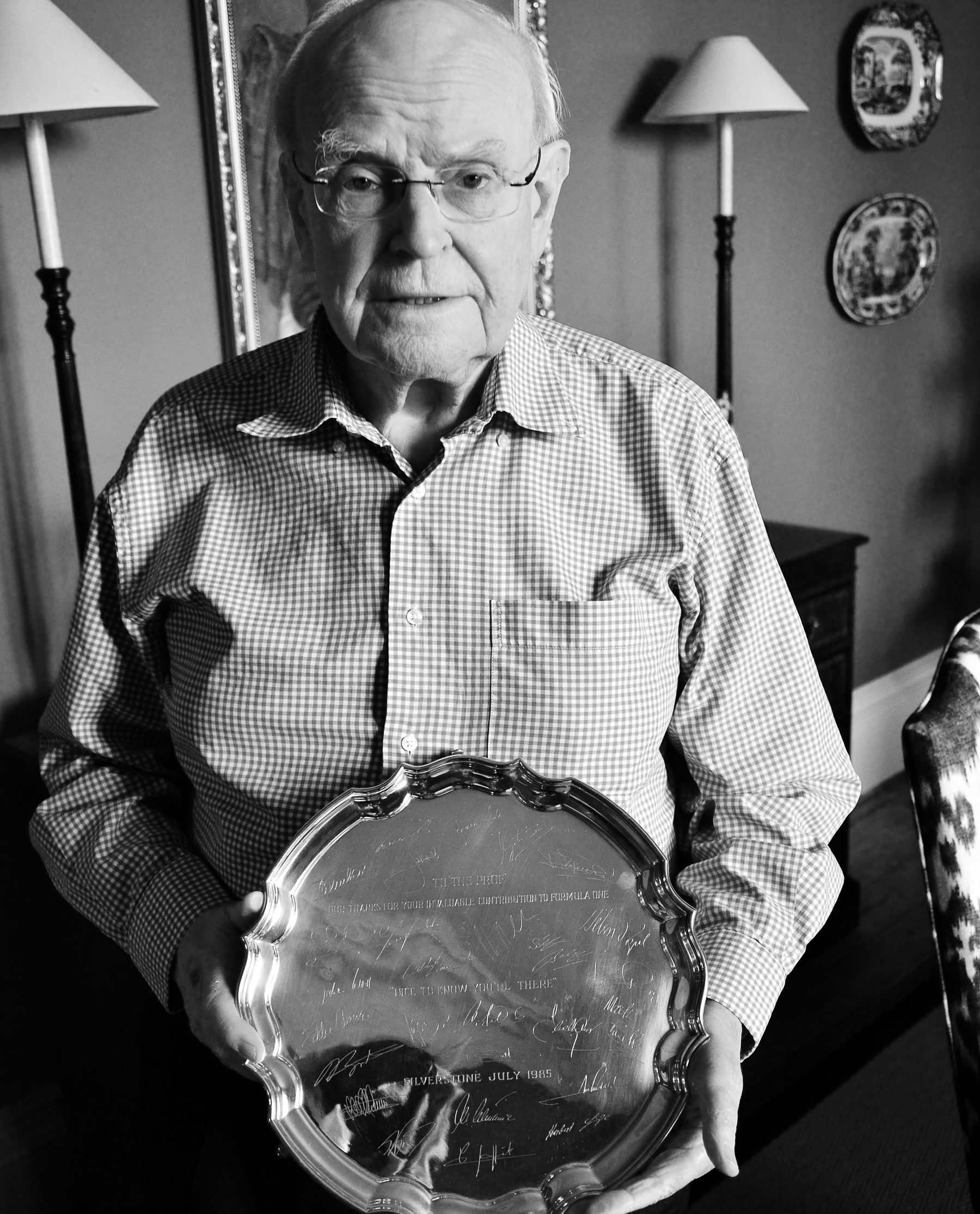 An elderly man with white hair and glasses, in a checked shirt, looks towards the camera in a room with lamps and plates on the wall.