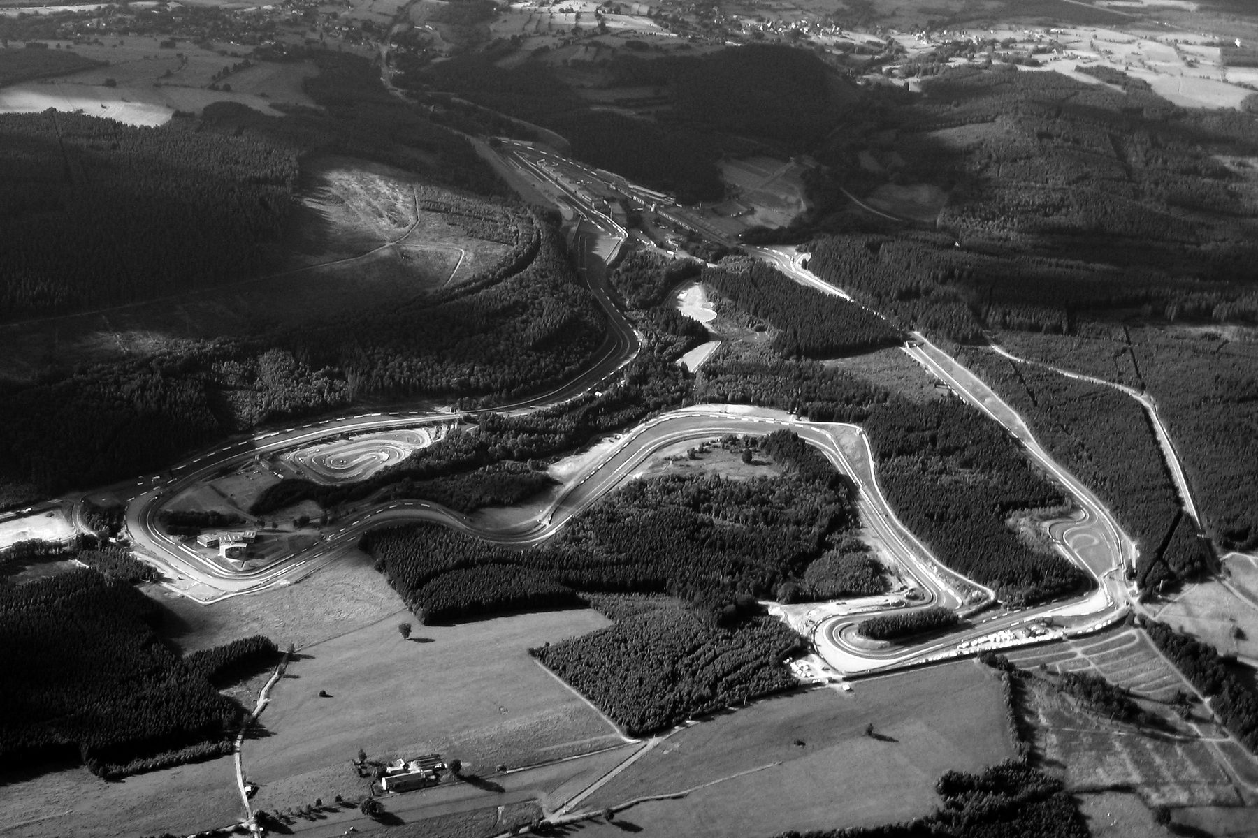 An aerial view over wooded hills and fields, the ribbon of a long racing circuit winding through the valley and up through the forest.