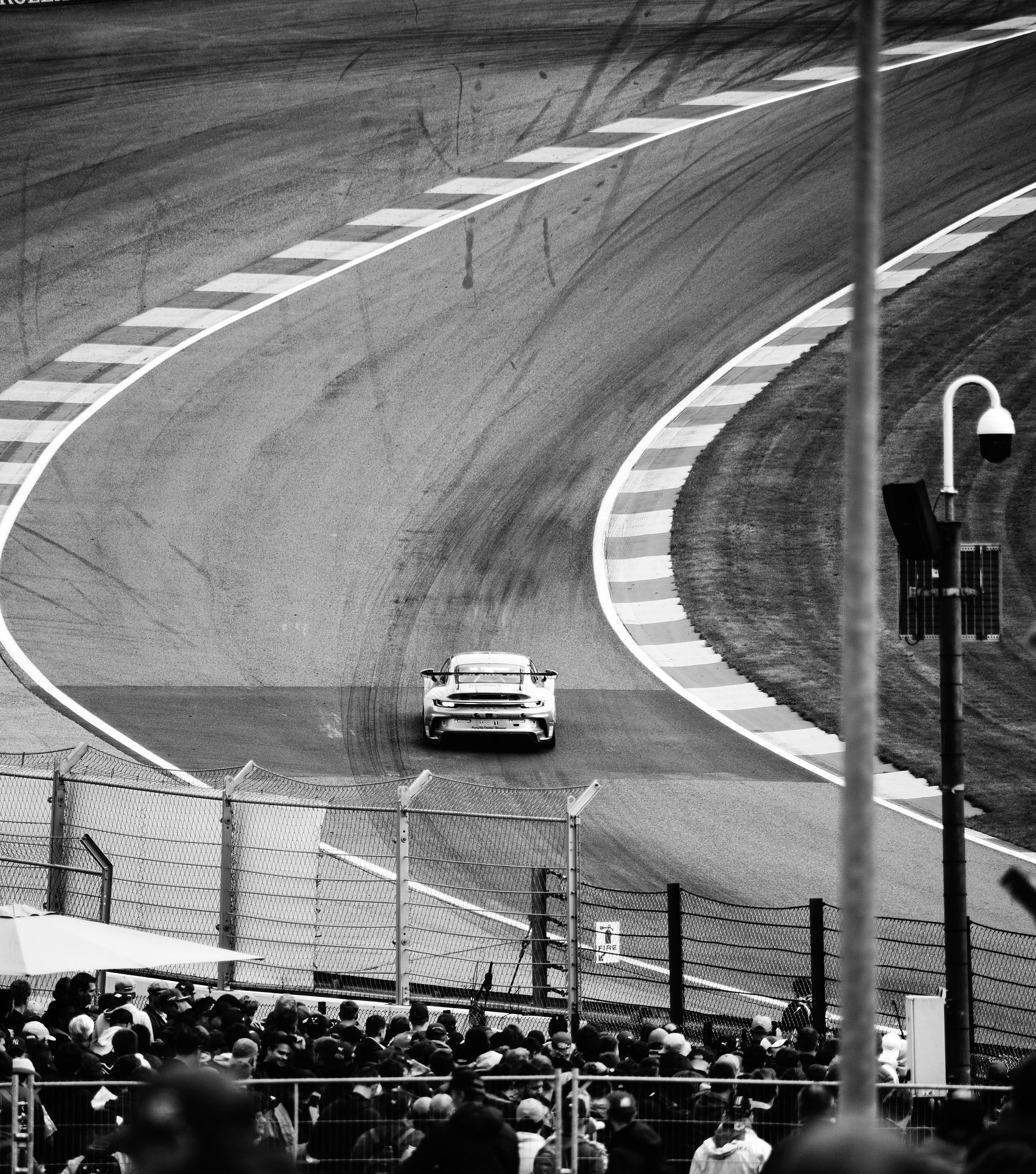 From high above, a single car climbs a steep left-right sweep of track between striped kerbs, spectators crowding the fence below.