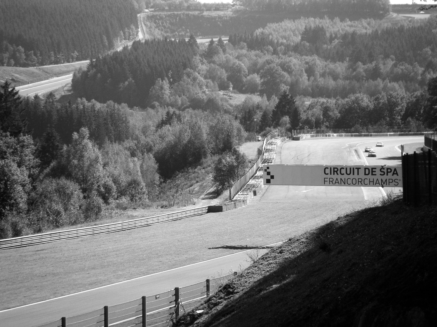 Vista ladera abajo hacia un valle de bosque espeso; el circuito sale de entre los árboles y sube hacia un edificio de boxes, con una pancarta de «Circuit de Spa-Francorchamps» sobre la pista.