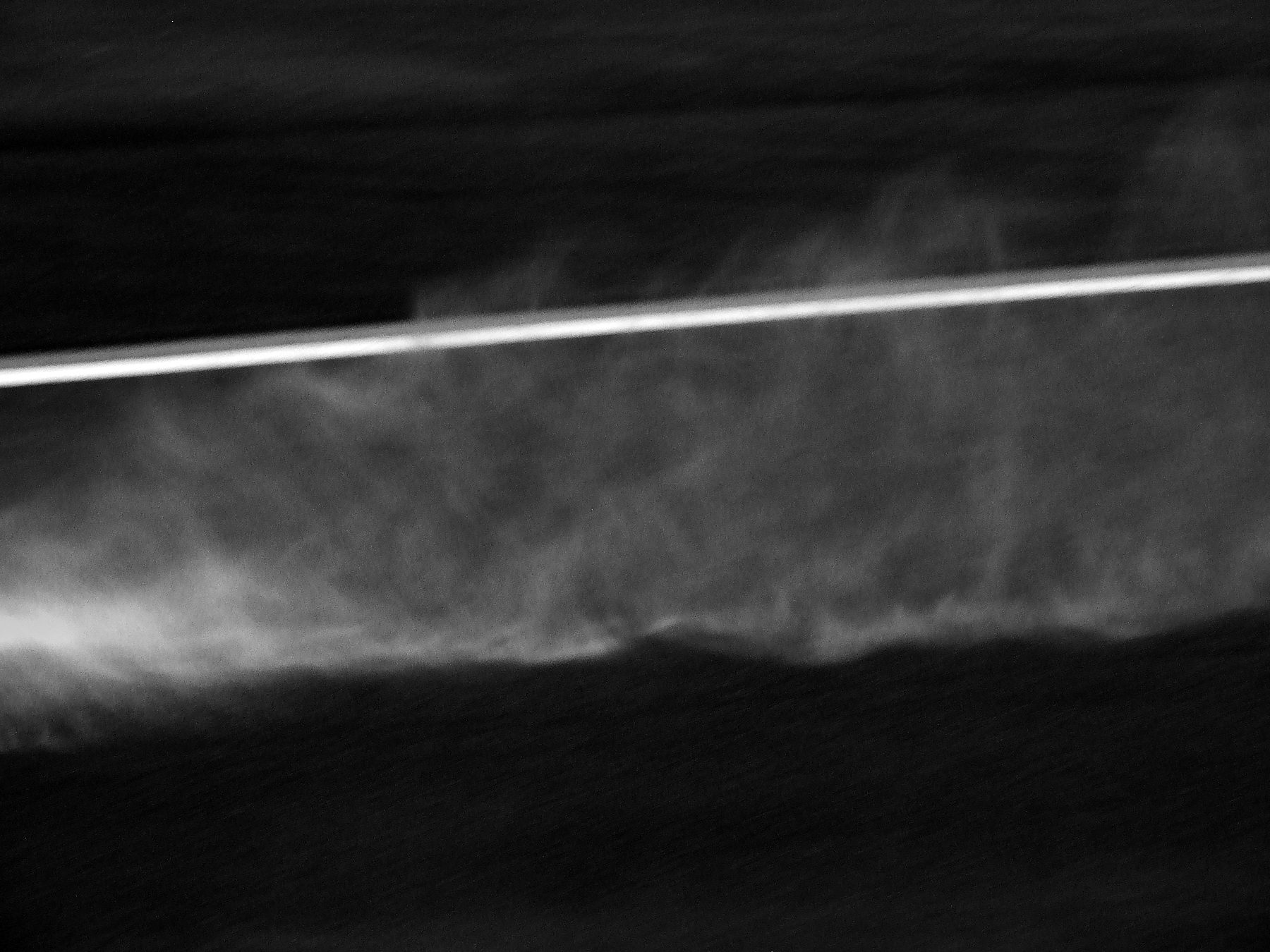An abstract streak of spray and mist across a dark frame, a light glowing through it.