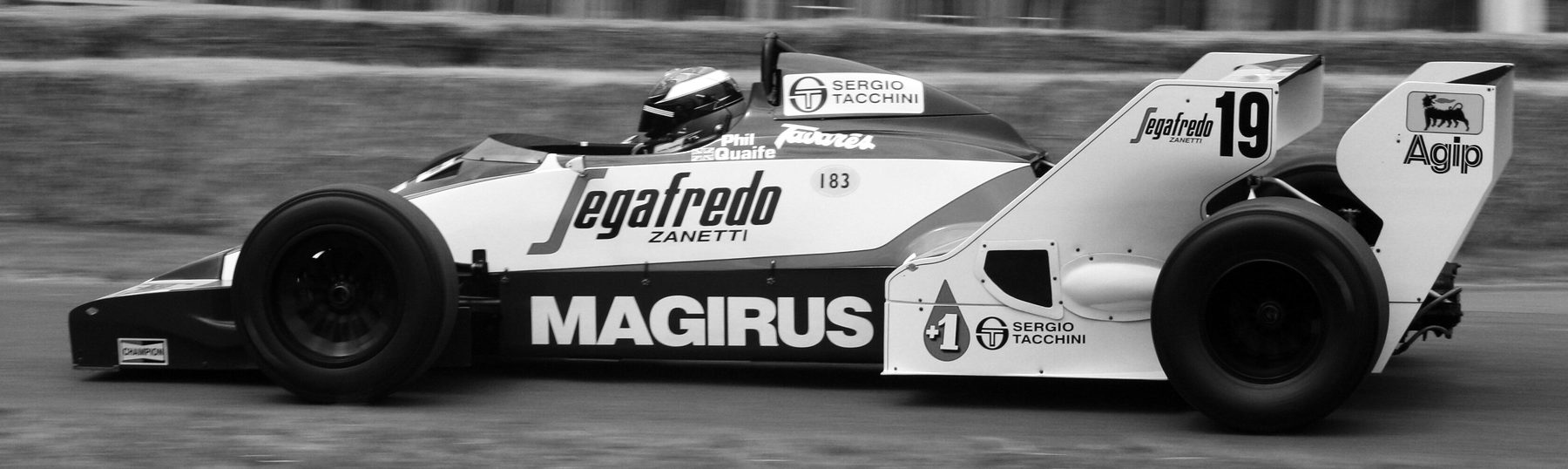A white single-seater numbered 19, lettered ‘Segafredo’ and ‘Magirus’, runs in profile, its driver’s helmet just above the cockpit, the background a blur.