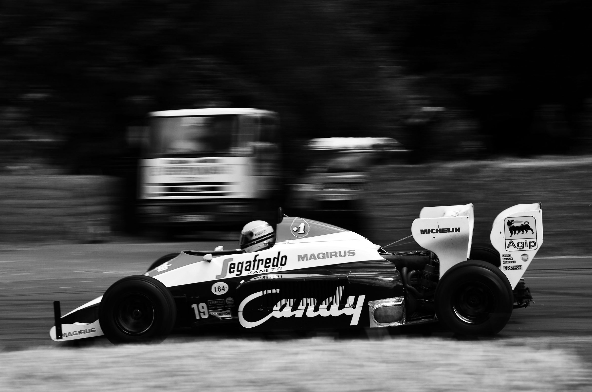 A white Toleman numbered 19, lettered ‘Candy’ and ‘Segafredo’, streaks past in a panning shot, the lorry and bank behind it a blur of speed.