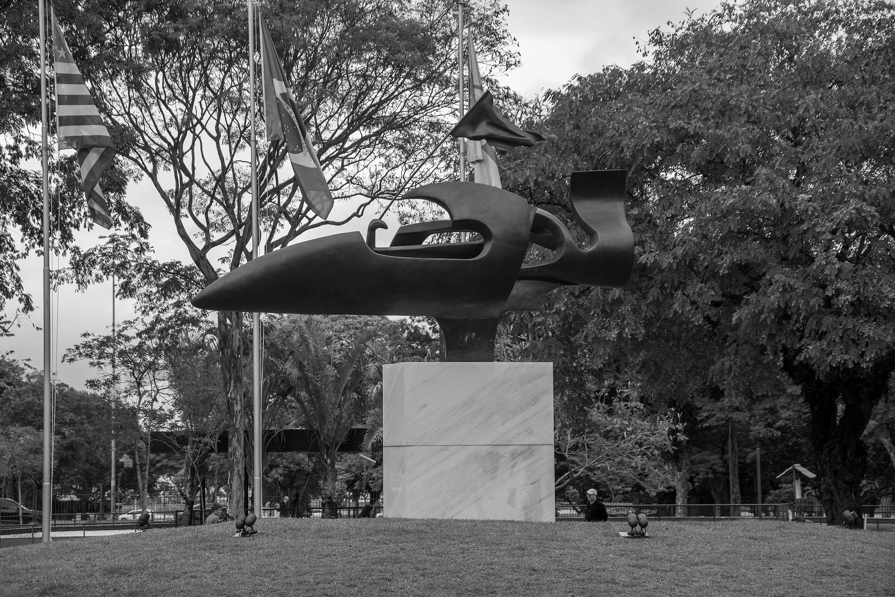 A bronze sculpture like the swept nose and cockpit of a racing car, mounted high on a white stone plinth among trees and flagpoles in a park.
