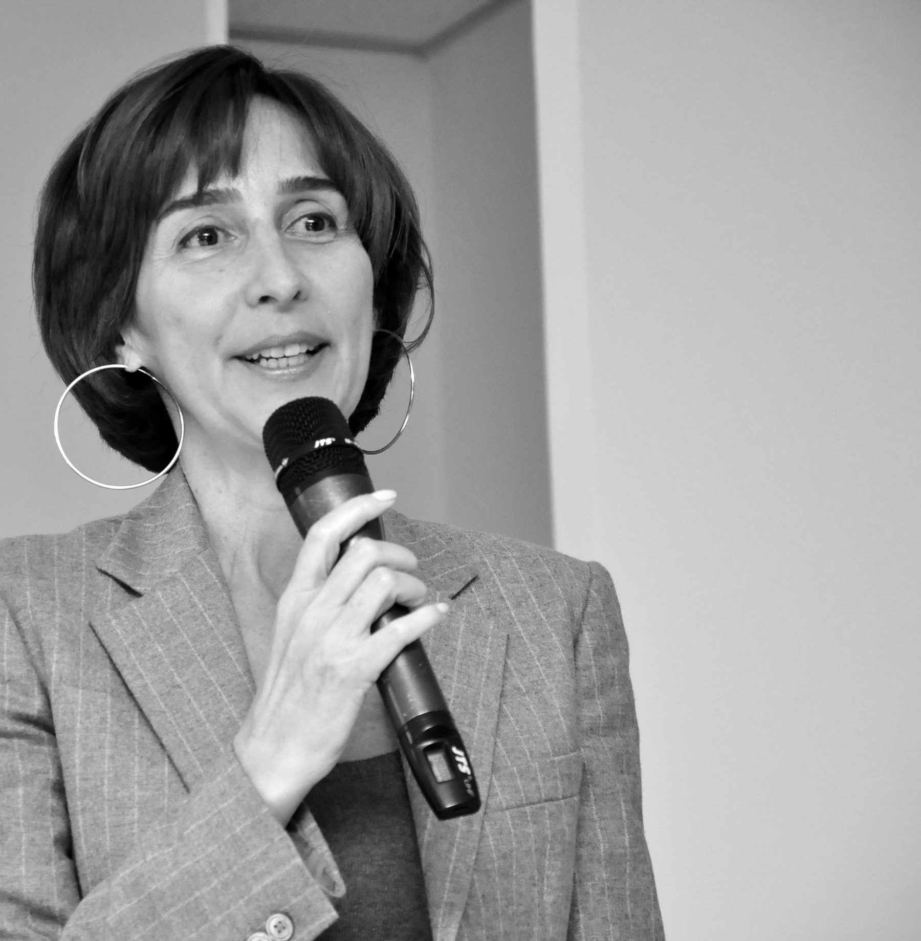 A woman with a dark bob and large hoop earrings smiles as she speaks into a hand microphone.
