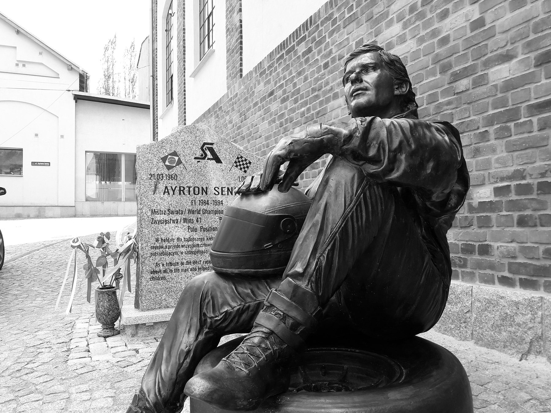 A life-size bronze of a racing driver sitting on a tyre, one arm resting on the helmet in his lap, gazing upwards, in front of a brick wall and an engraved memorial stone.