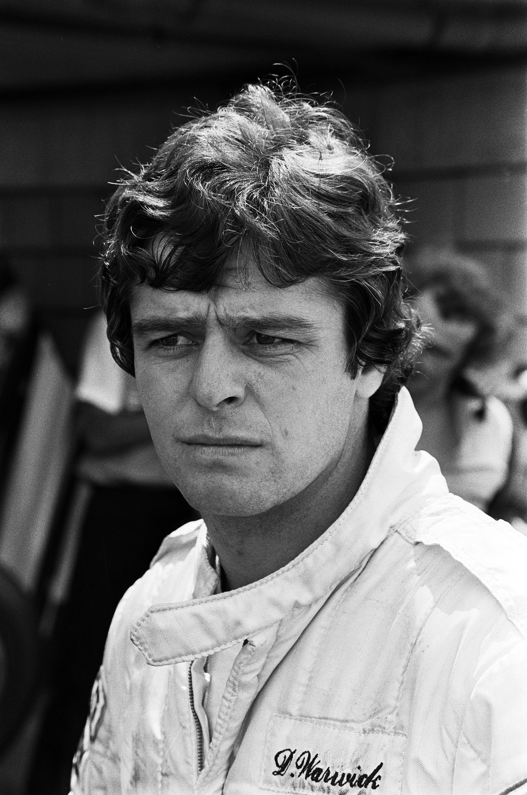 Portrait of a young driver with thick, tousled hair in white overalls, looking to one side with a serious expression.