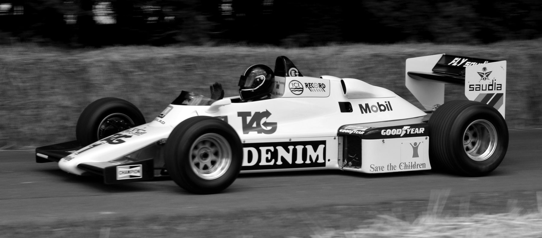 A white Formula One car lettered TAG, Denim and Mobil runs side-on past straw bales, dark woodland blurred behind it, its driver in a black helmet raising a hand.