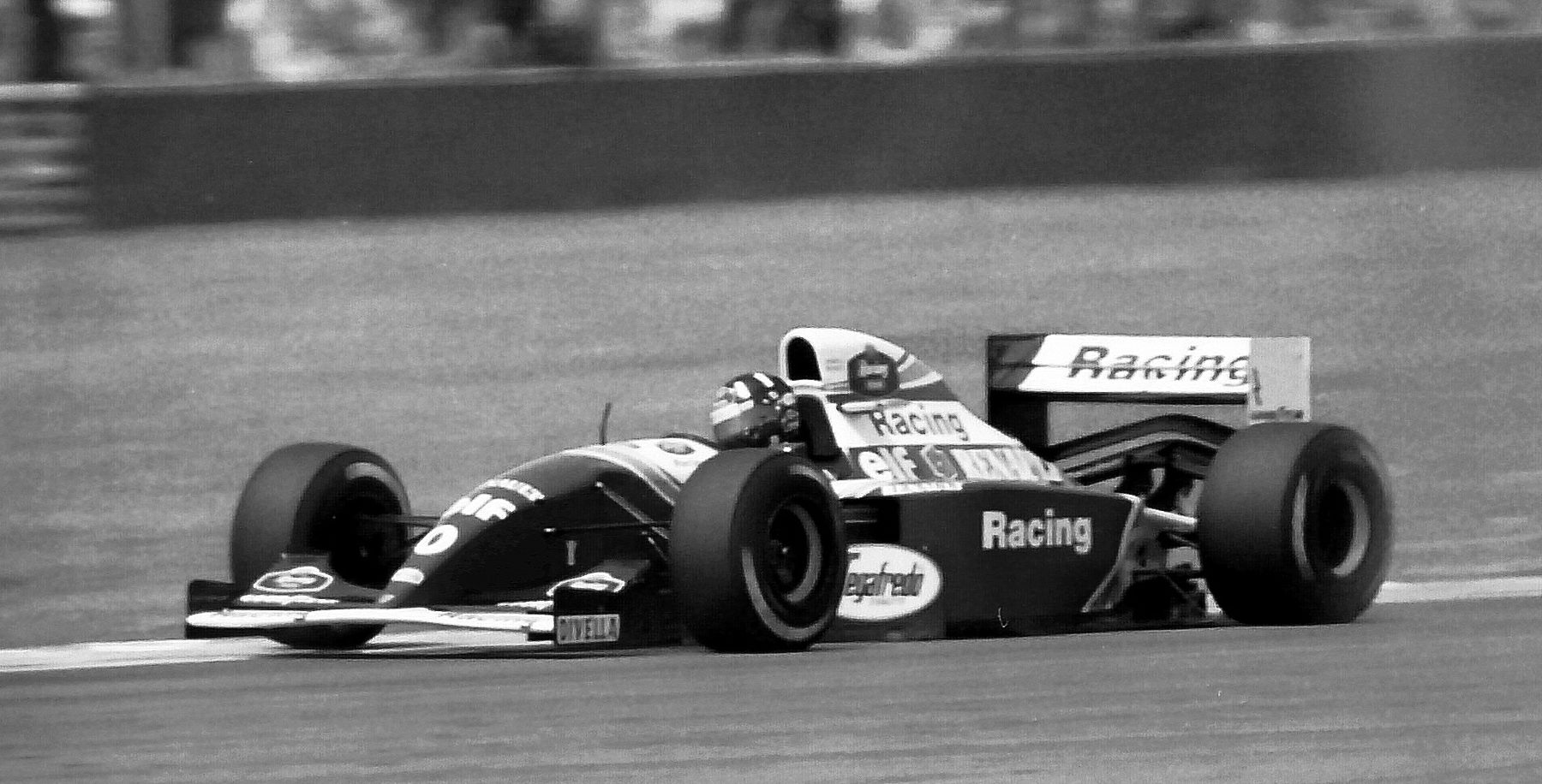 A dark Williams with pale lettering, ‘Racing’ on its sidepod and rear wing, runs along a straight past a grass verge.