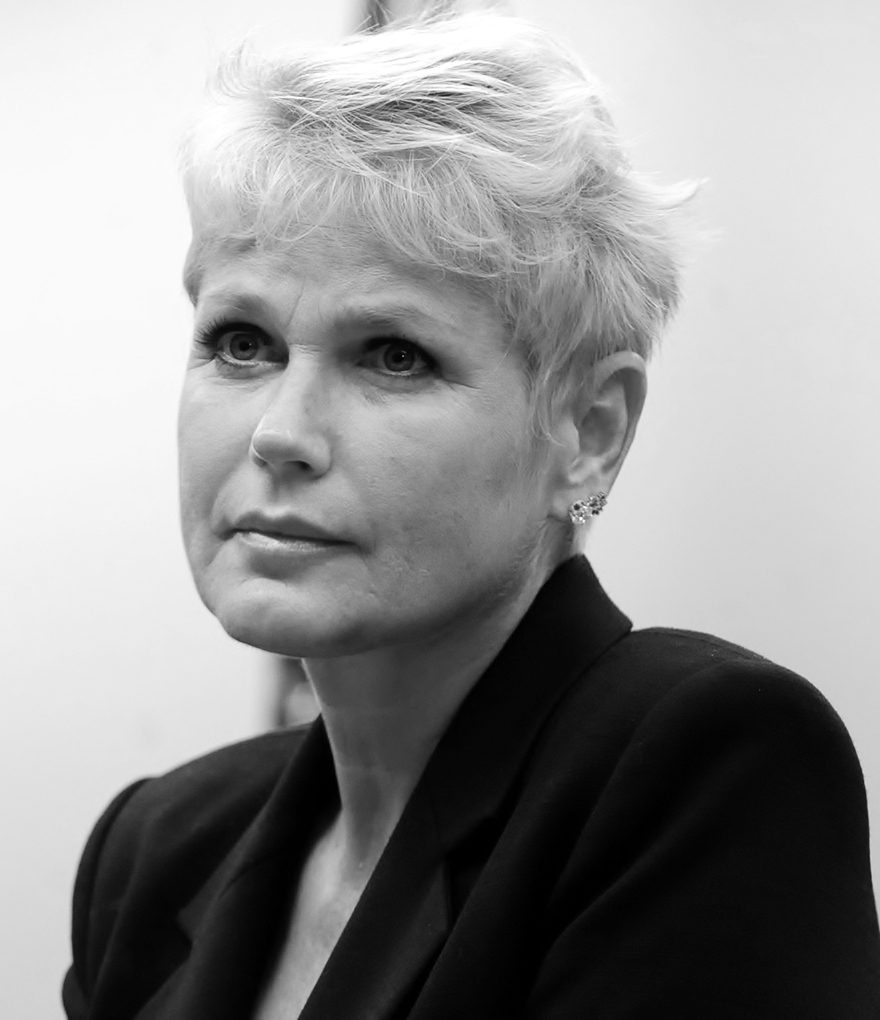 A woman with short, tousled, very pale hair, in a black jacket, looks off to one side, listening.
